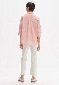 Camicia rosa chiaro con una piega sul retro, maniche a tre quarti e colletto con bottoni, abbinata a pantaloni bianchi e scarpe da ginnastica chiare.