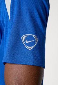 Blå atletisk skjorte med struktureret stof, der har et hvidt broderet Nike-logo på ærmet. Nærbillede af skulderområdet.