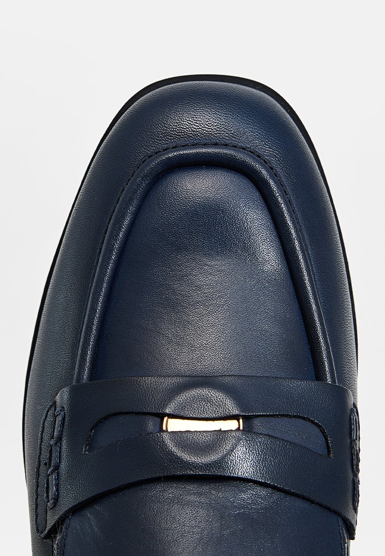 Mocassino in pelle blu navy con punta arrotondata, texture liscia e accentuazione in metallo dorato sulla striscia. Design minimalista, linea classica.