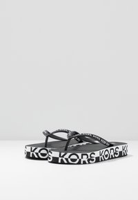 Mustat flip-flopit, joissa on kumiset hihnat, joissa toistuu valkoinen "KORS" -teksti, ja teksturoitu musta pohja. Litteä muotoilu ilman lisäkoristeita.