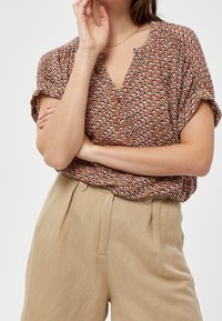 Blusa marrone a fantasia con scollo a V e maniche corte, abbinata a pantaloni beige a vita alta. Il tessuto sembra leggero e morbido.