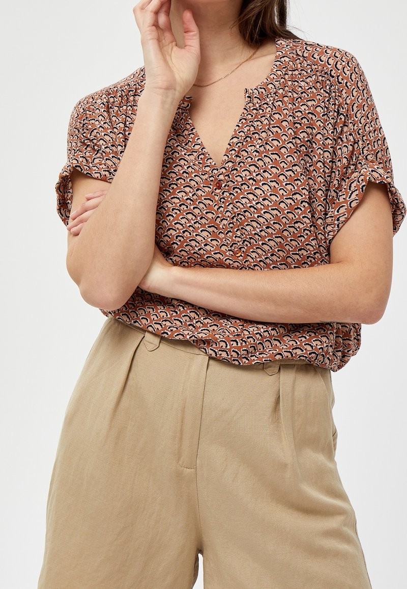 Blusa marrone a fantasia con scollo a V e maniche corte, abbinata a pantaloni beige a vita alta. Il tessuto sembra leggero e morbido.