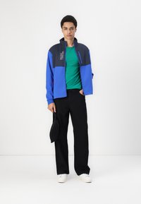 Modra in siva zip-up jakna z visokim ovratnikom, kombinirana z zeleno majico, črnimi hlačami in belimi supergami. Drži črno torbico.