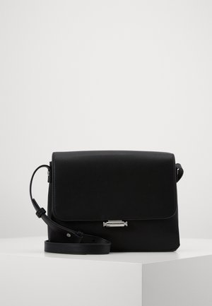Bolso bandolera de cuero negro con cierre de solapa y broche rectangular plateado, correa ajustable colocada sobre un pedestal blanco contra un fondo liso.