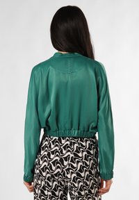 Teal satin bluse med krave og elastisk talje, parret med bukser med sort-hvidt spiral mønster, set fra ryggen.