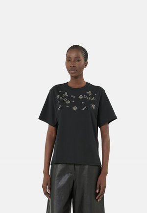 Donna che indossa una maglietta nera a maniche corte con decorazioni floreali argento sul petto e pantaloni in pelle neri, in piedi su sfondo bianco.