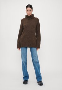 Brauner, übergroßer Strickpullover mit Rollkragen und gerippten Bündchen, kombiniert mit hellblauen geraden Jeans und schwarzen Sneakers.