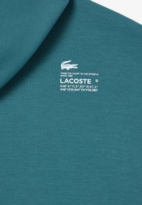 Tissu teal avec le logo blanc de Lacoste, emblème de crocodile, slogan "Du court aux rues depuis 1933" et deux ensembles de coordonnées géographiques.