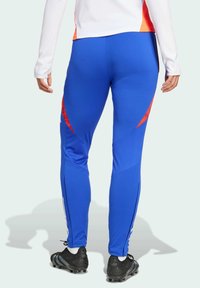 Blaue sportliche Leggings mit roten und strukturierten Akzenten, aus einem glatten Stoff gefertigt, mit Reißverschlüssen an den Knöcheln und schwarzen Schuhen.