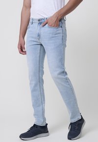 Jeans de denim azul claro con corte ajustado, ligeros desgastes y detalles de costura. Llevados con zapatillas deportivas oscuras.