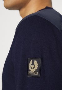 Námořnicky modrý pletený svetr s texturovanou tkaninou; má tmavě modrý panel na rameni a černou nášivku s logem zdobeným zlatem na rukávu.