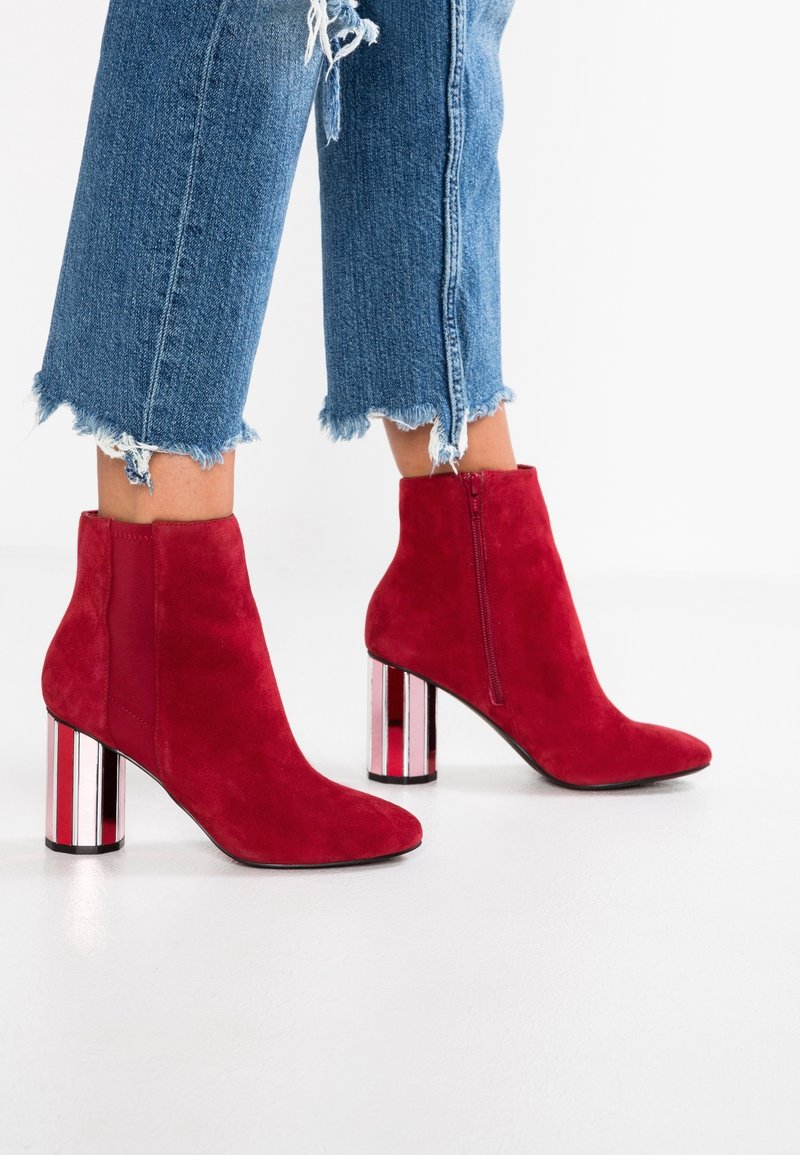 Bottes chevilles en daim rouge avec un bout pointu et une fermeture éclair latérale. Dotées d'un talon épais miroir avec des rayures verticales. Portées avec un jean effiloché.
