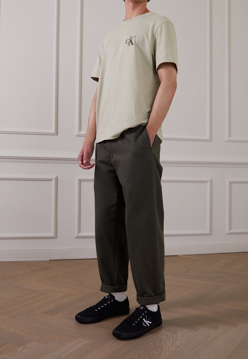 Personne portant un t-shirt beige Calvin Klein, un pantalon vert olive avec revers, des chaussettes blanches et des baskets Calvin Klein noires, debout à l'intérieur.