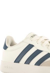 White sneaker with navy blue stripes, leather upper, textured heel, and lace-up front. Features embossed logo on the side.