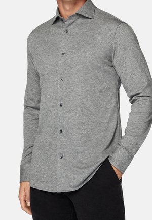 Mand iført en grå langærmet skjorte med knapper og krave, venstre hånd i lomme på sorte bukser foran en ensfarvet baggrund.