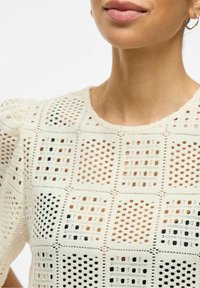 Närbild av en kvinna som bär en krämfärgad virkadräkt med fyrkantiga mönster och guldiga hoopsörhängen mot en vit bakgrund.