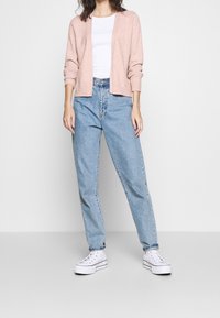Cárdigan rosa claro, con puños acanalados y frente abierto. Camiseta blanca debajo. Jeans de tiro alto en azul claro. Zapatillas de plataforma blancas con rayas negras.