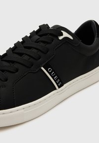 Svart lær sneaker med flate snørebånd, hvit gummisåle og et svart-hvitt logo bånd langs siden. Glatt tekstur, enkelt design.