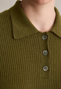 Polo a coste verde oliva con colletto e tre bottoni. Il tessuto ha una consistenza morbida e una lucentezza sottile.
