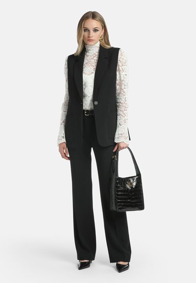 Zwarte op maat gemaakte vest over een witte kanten top met lange mouwen, gecombineerd met zwarte broek en pointy heels. Heeft een glanzende zwarte handtas.