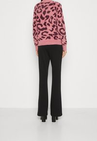 Personne portant un pull duveteux rose à imprimé léopard, un pantalon noir à jambes larges et des bottes noires à talons, se tenant devant un fond uni.