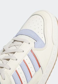 Zapatilla de cuero blanca con rayas en zigzag de color azul claro y rojo, con detalles perforados, acentos en gris claro y cierre de cordones beige.