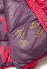 Intérieur de la veste matelassée en violet avec une surface texturée. Présente des icônes jaunes indiquant la résistance thermique, à l'eau et au vent, ainsi que l'utilisation de fibres recyclées.