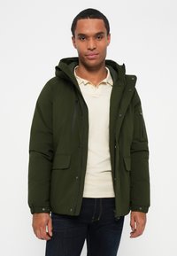 Giacca imbottita verde oliva con cappuccio, chiusura con zip e molteplici tasche. Presenta polsini elasticizzati e una superficie liscia e opaca.