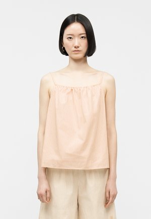 Jeune femme aux cheveux courts noirs portant un haut sans manches ample rayé pêche et blanc ainsi qu'un pantalon ample assorti, debout de manière neutre.