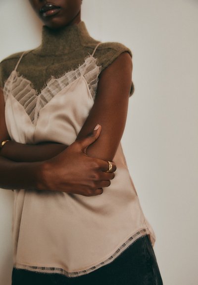 Beige satin camisole with sheer ruffled trim, layered over olive-green knit top. Model's hands clasped, wearing a gold ring.
