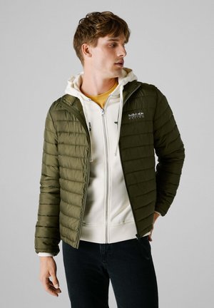 Olive green puffer jacket with horizontal stitching, front zipper, and logo on left chest, worn over a white hoodie and yellow shirt.