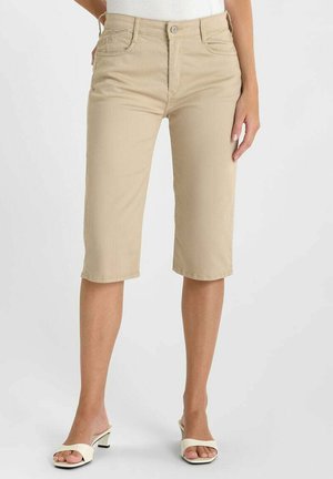 Vrouw draagt beige knielange shorts en witte open-teen sandalen met hak, staand tegen een effen achtergrond.