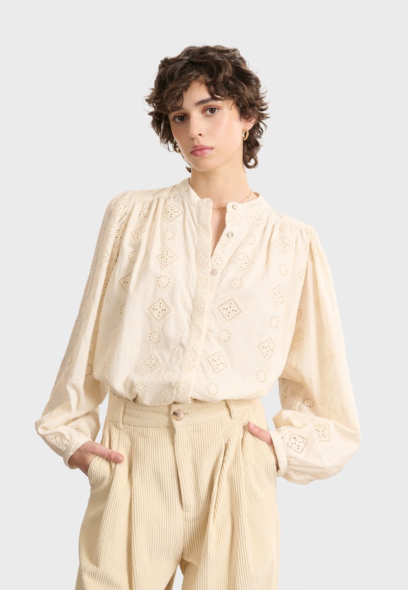 Crème-kleurige blouse met geborduurde diamanten en bloemenpatronen, knoopkraag en puffed lange mouwen. Gecombineerd met een structuurbeige broek.