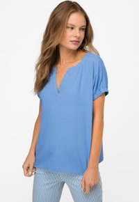 Top azzurro chiaro a maniche corte con scollatura a V e risvolti. Realizzato in un tessuto morbido. Abbinato a pantaloni a righe blu e bianchi.