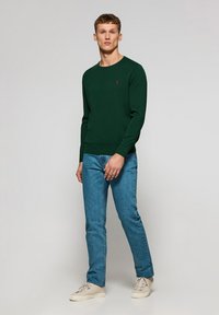 Grøn langærmet sweater med rund halsudskæring, ribbede manchetter og kant, parret med lyseblå jeans og hvide sneakers. Rød logo på brystet.