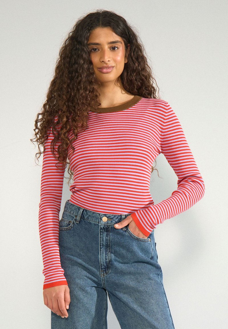 Jeune femme aux longs cheveux bouclés portant un haut à manches longues rayé rouge et blanc et un jean taille haute bleu, debout devant un fond uni.