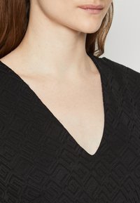 Tissu noir avec un motif géométrique texturé, présentant un design en V. Le matériau semble doux et léger.