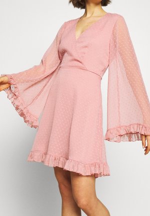 Femme portant une robe rose à pois avec des manches longues en forme de cloche en tissu transparent et des ourlets à volants, debout devant un fond uni.