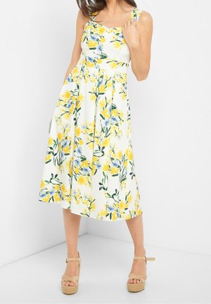 Kvinde iført en ærmeløs hvid midikjole med gul og blå blomsterprint og beige platformsandaler, stående op ad en hvid væg.