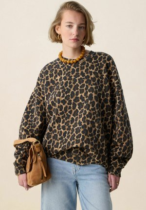 Femme portant un haut à manches longues imprimé léopard, un jean bleu clair, un collier de perles ambrées, des boucles d'oreilles créoles en or, tenant un sac à main en daim marron.