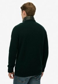 Dunkelgrüner Pullover mit strukturiertem Finish, mit einem umgeschlagenen Kragen im Karomuster. Getragen über blauen Jeans, was einen lässigen Look zeigt.