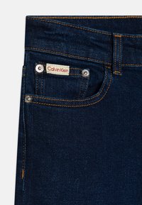 Jeans in denim blu scuro con una tasca frontale con etichetta "Calvin Klein" bianca e rossa, accessori in argento e cuciture arancioni a contrasto.