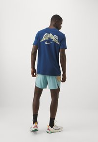 Tumši zils Nike T-krekls ar kalna grafiku uz muguras, saskaņots ar gaiši zilām šortiem ar sandalkrāsas akcentiem. Sporta apavi ar baltām un zaļām detaļām.