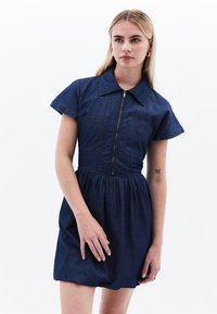Denimmekko, jossa on vetoketjukiinnitys edessä, lyhyet hihat ja kaksi rintataskua. Mekossa on istuva vartalo ja levenevä hame syvässä sinisessä.