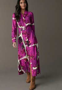 Rochie cu mâneci lungi, cu nasturi, într-un violet vibrant cu modele florale și accente albii. Are o croială lejeră și fante laterale. O pereche de bocanci maronii.