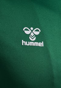 Zielona sportowa koszulka wykonana z gładkiej tkaniny, z białym logo o geometrycznych wzorach oraz napisem „hummel” poniżej.