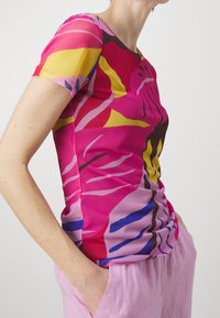 Henkilö, joka on pukeutunut sopivan kokoiseen lyhythihaiseen paitaan, jossa on kirkkaat abstraktit vaaleanpunaiset, keltaiset, siniset ja mustat kuviot, sekä vaaleanpunaisiin housuihin, käsivarret taskuissa.