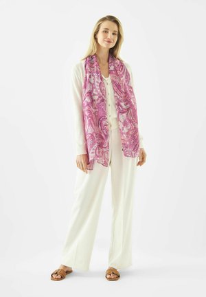 Vrouw draagt een witte cardigan met knopen en broek met wijde pijpen, gecombineerd met een roze sjaal met patroon en bruine sandalen met open tenen.