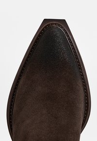 Zapato de ante marrón con punta puntiaguda, que presenta un acabado suave y costuras de un marrón oscuro en contraste a lo largo del borde.