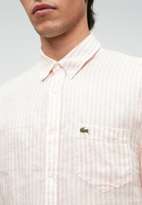 Ροζ ανοιχτό ριγέ κοντομάνικο πουκάμισο από ελαφρύ ύφασμα, με γιακά με κουμπιά και μικρό πράσινο λογότυπο στην τσέπη.